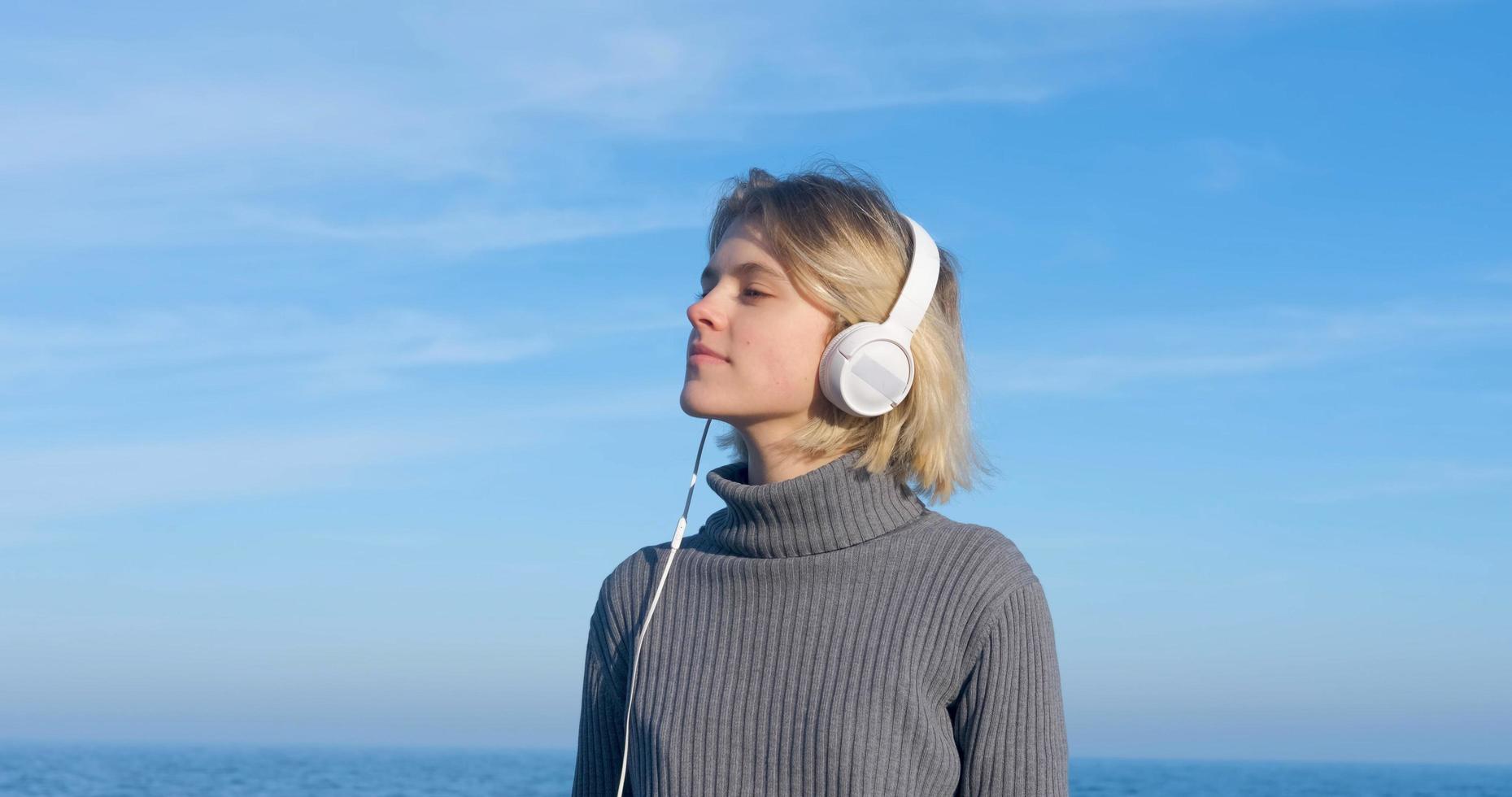 jonge knappe vrouw luistert naar muziek met een koptelefoon buiten op het strand tegen de zonnige blauwe hemel foto