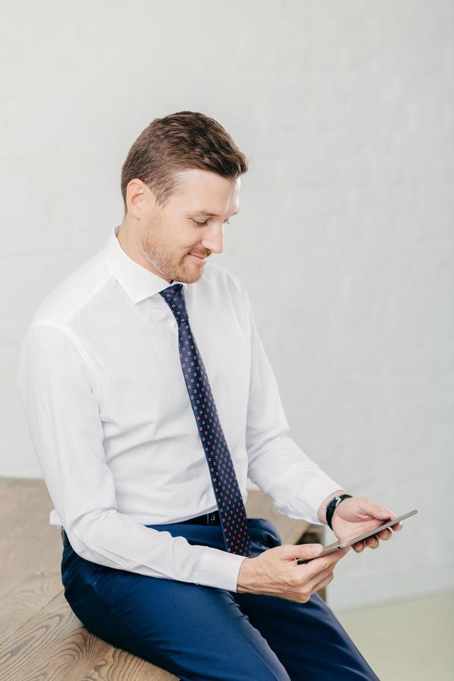 jonge ongeschoren intelligente man draagt formele kleding, gebruikt digitale tablet voor werk, gefocust op scherm, zit aan bureau, geïsoleerd op witte achtergrond. programmeur ontwikkelt applicatiesoftware foto