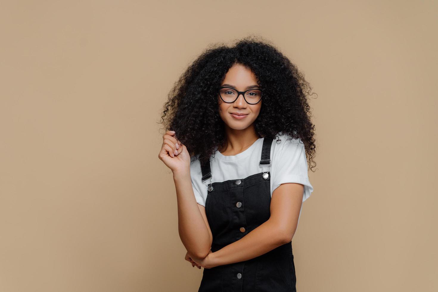 geïsoleerd schot van aangenaam ogende jonge afro-amerikaanse vrouw raakt krullend haar aan, heeft borstelig kapsel, draagt een optische bril, wit t-shirt en zwarte overall, poseert over bruine studioachtergrond foto