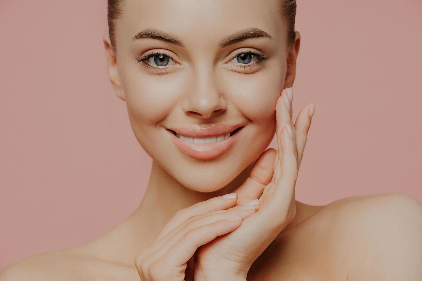 close-up shot van mooie vrouw met blote schouders, raakt het gezicht zachtjes aan, heeft een gezonde huid, natuurlijke make-up, kijkt direct naar de camera, geïsoleerd op roze achtergrond. schoonheid, gezichtsverzorging en spa-concept foto