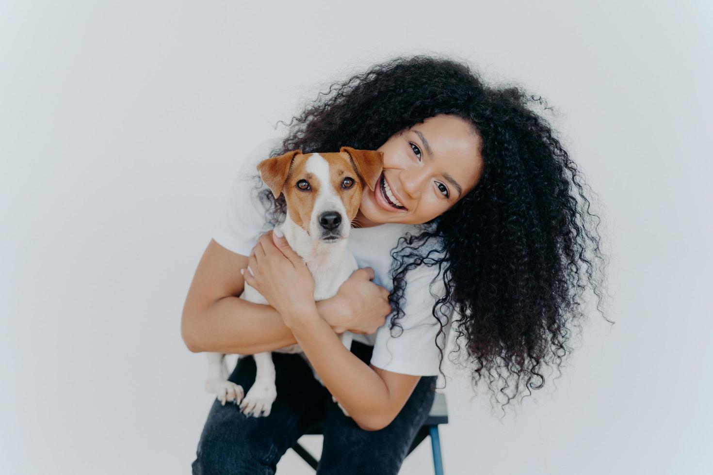 gelukkige gekrulde vrouw kantelt het hoofd, houdt rashond vast, heeft een vrolijke uitdrukking, glimlacht aangenaam, heeft een krullend kapsel, draagt een wit t-shirt, geïsoleerd. afro dame aaien favoriete huisdier. foto