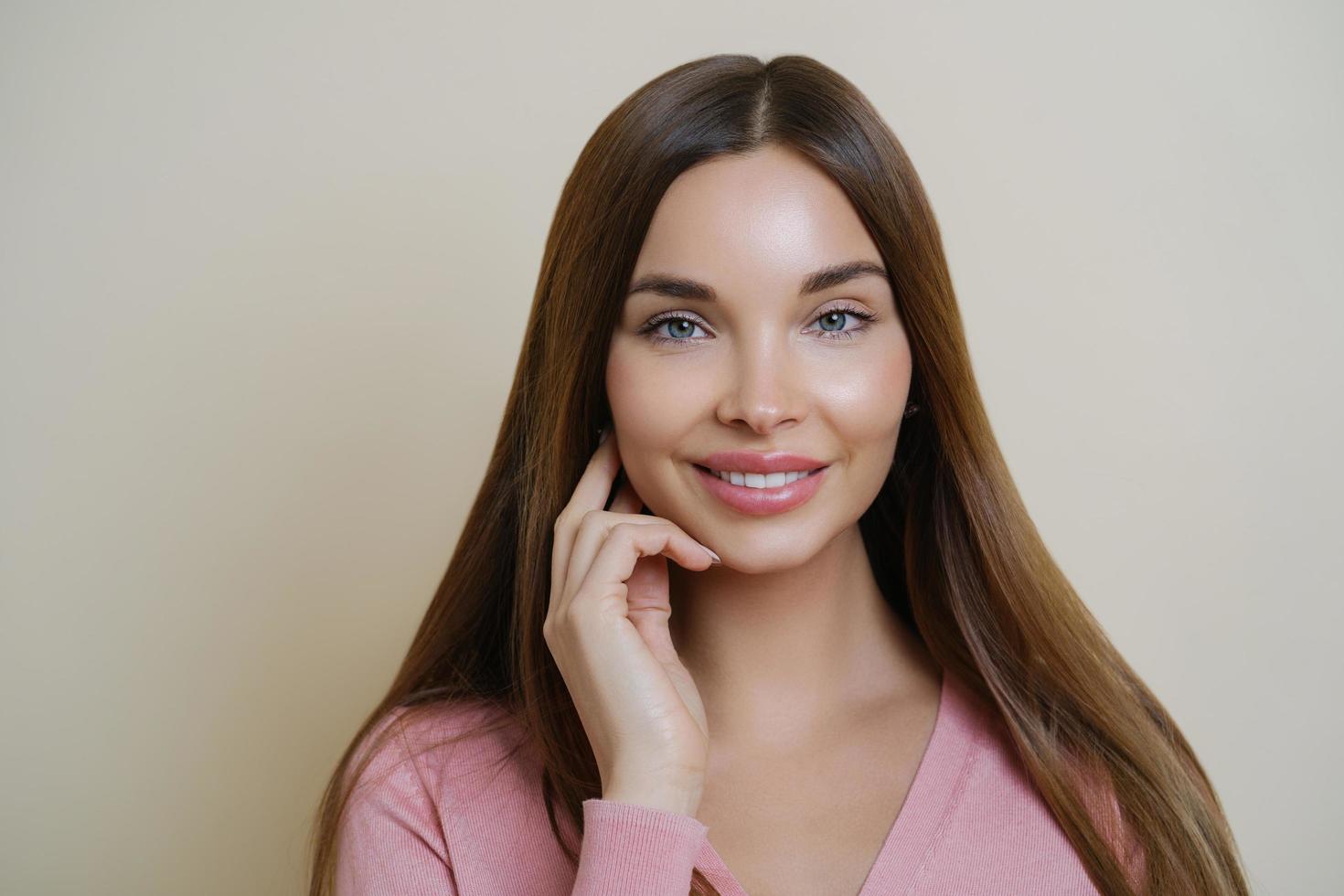 schattig jong vrouwelijk model lacht aangenaam, heeft lang donker haar, kijkt direct naar de camera met tevreden uitdrukking, heeft een perfect schone huid, draagt een roze trui, geïsoleerd op beige achtergrond foto