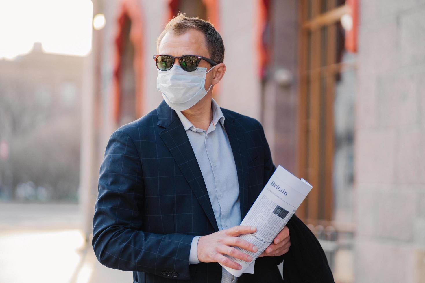 zakenman draagt beschermend masker tegen overdraagbare besmettelijke ziekte, leest kranten, is elegant gekleed, poseert buiten, denkt na hoe nieuw coronavirus uit china te voorkomen. griep in de stad. foto