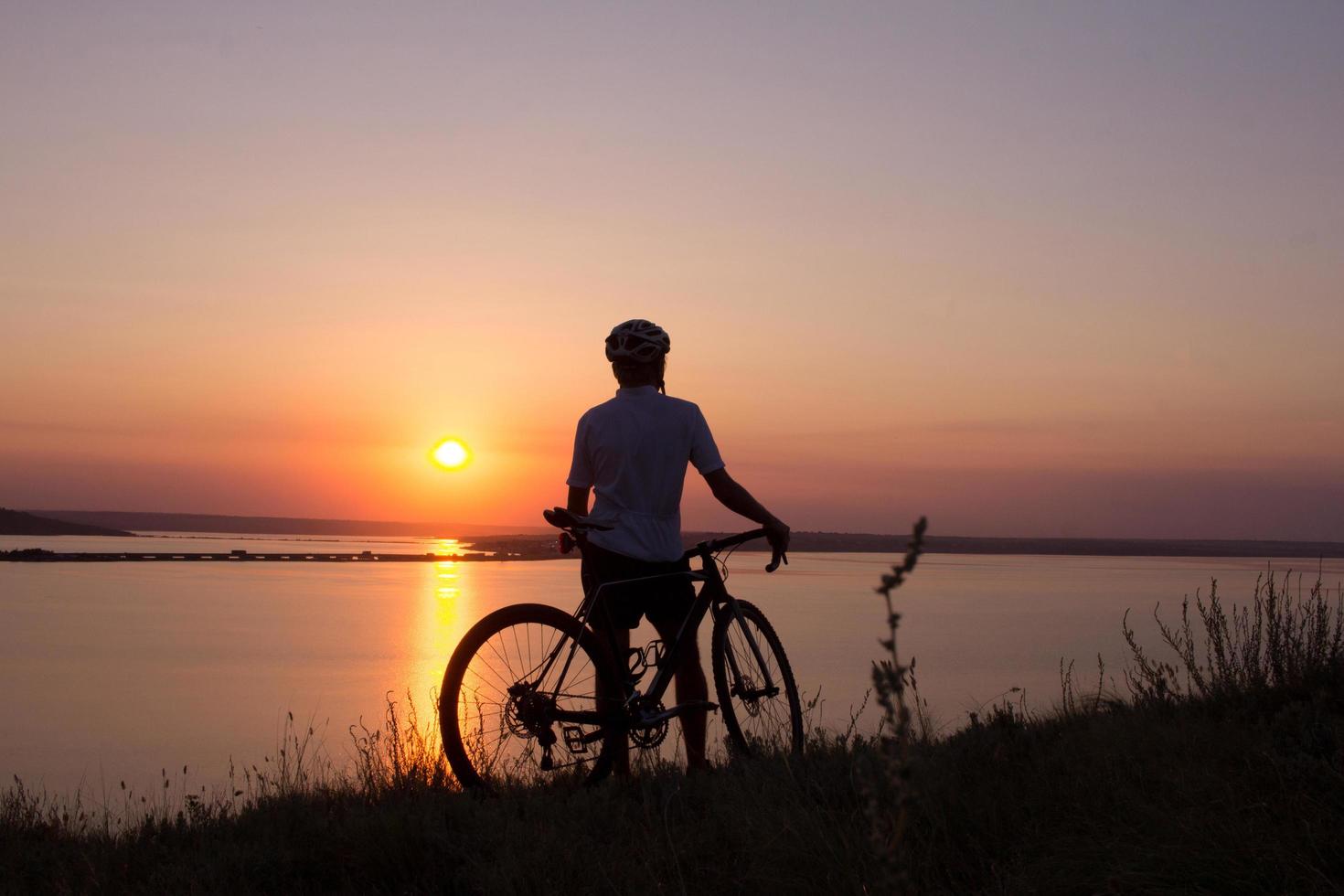 silhouet van een fietser die naar zonsondergang in het meer kijkt, mannelijke fietser in helm tijdens zonsondergang foto