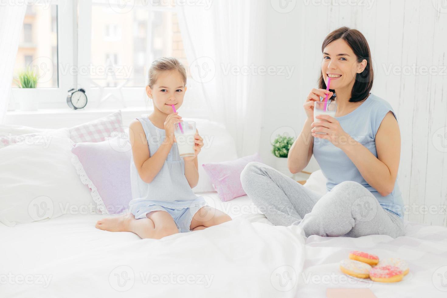 vrolijke moeder en dochter gekleed in pyjama, ontbijten in de ochtend, milkshake drinken met donuts, gekruiste benen zitten op comfortabel bed in witte slaapkamer. mensen en levensstijl concept foto