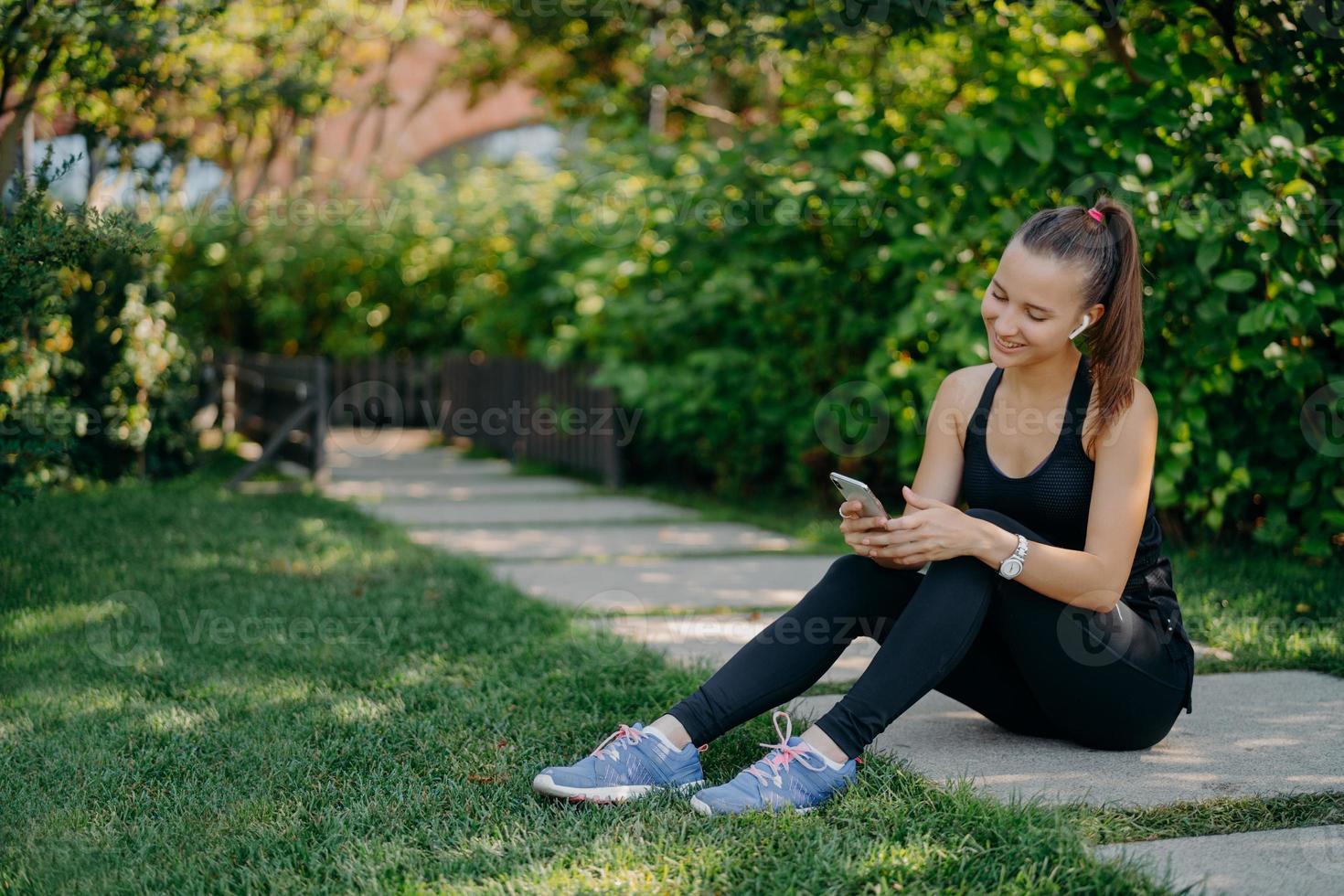 tevreden atletische vrouw in sportkleding lacht vrolijk surft op mobiele telefoon terwijl ze naar muziek luistert via draadloze koptelefoon neemt pauze na een zonnige ochtendtraining gebruikt app voor het volgen van activiteiten foto