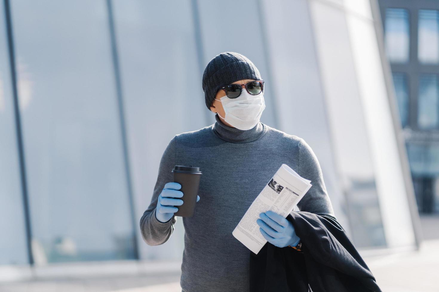 afbeelding van jonge man draagt steriel masker en rubberen medische handschoenen, kijkt opzij, loopt door de stad tijdens verspreiding van besmettelijke ziekten, drinkt koffie, opgevouwen krant. coronavirus preventie concept foto