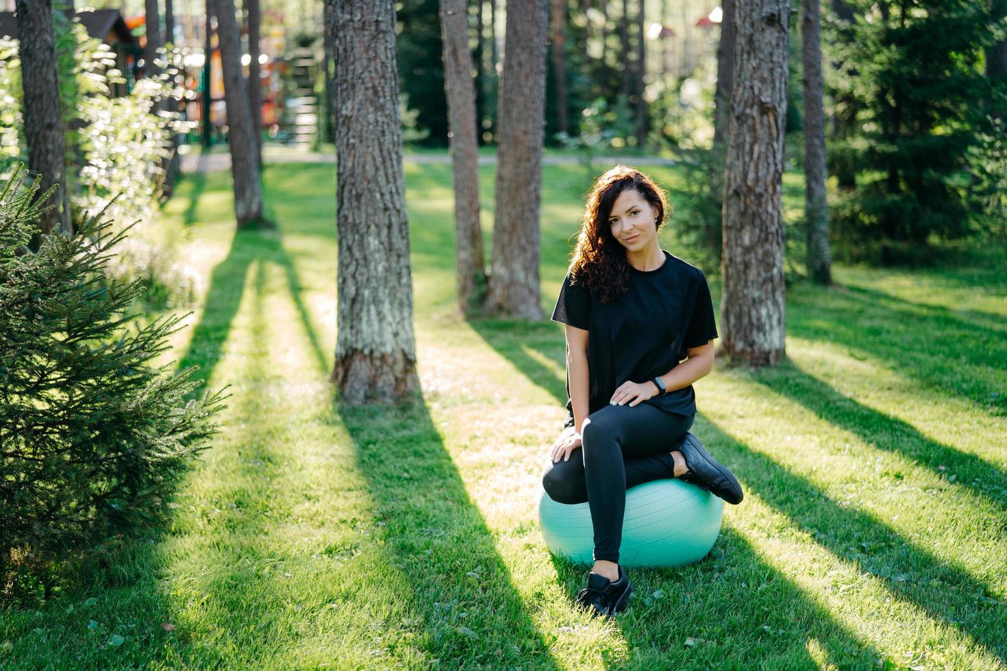 fit brunette jonge vrouw rust na het doen van oefeningen op fitness bal leidt actieve sportieve levensstijl poses in stedelijke omgeving op groen gras tegen bomen met zonneschijn doet gymnastiek. foto