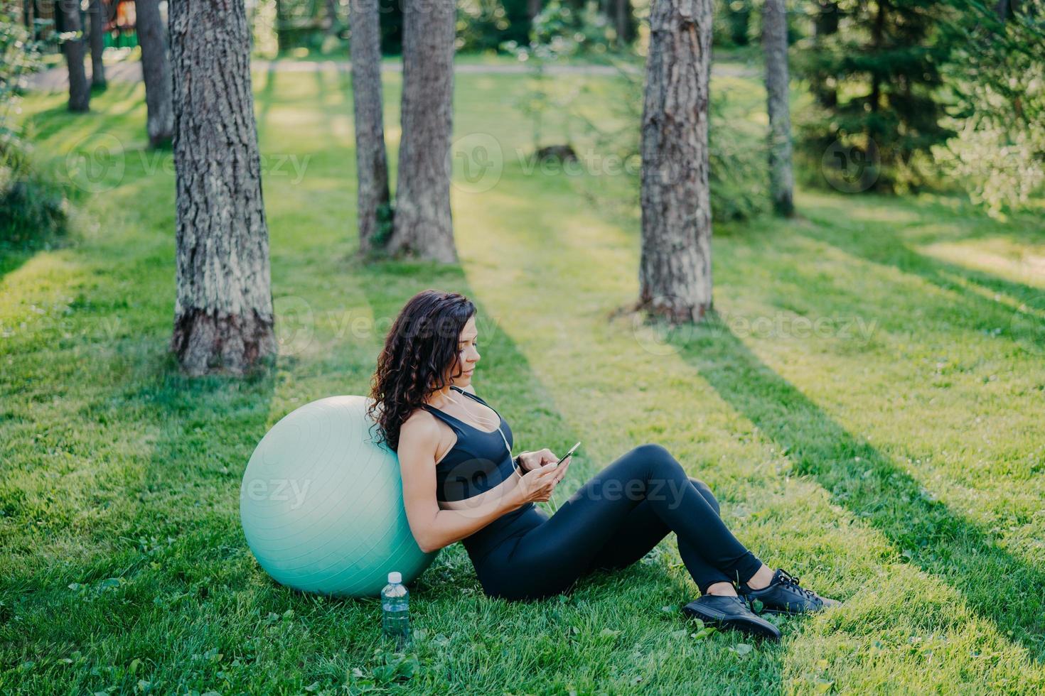 buitenopname van ontspannen brunette vrouw zit in de buurt van fitnessbal, gebruikt moderne mobiele telefoon, luistert naar muziek via oortelefoons, poseert op groen gras met een fles water, draagt sportkleding en sneakers foto