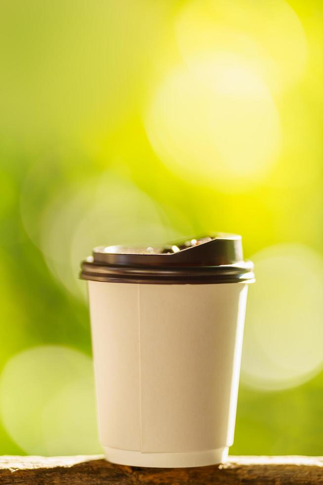 close-up van wegwerp papieren kopje warme koffie op houten balk met groene natuur achtergrond. foto