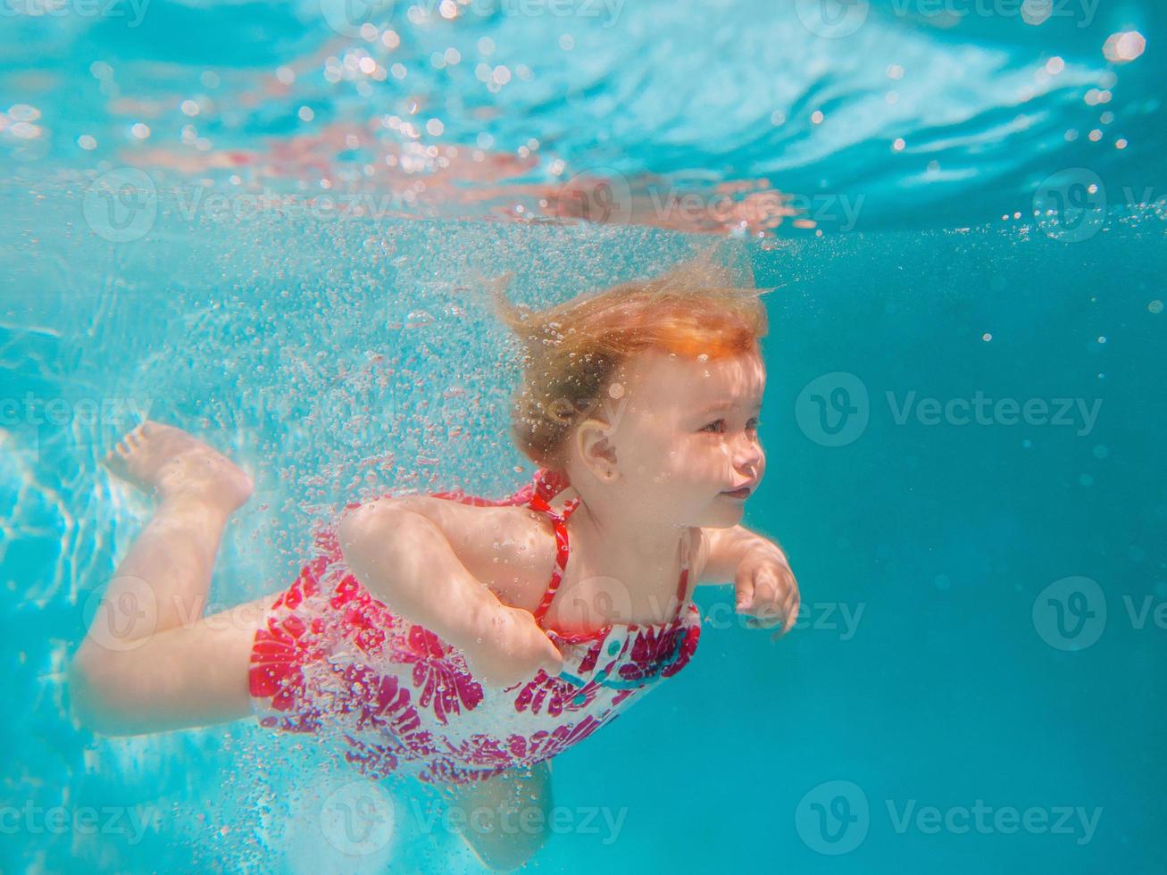 lachende babymeisje in schattige moderne jurk onderwater duiken in blauwe zwembad. actieve levensstijl, zwemles voor kinderen met ouders. watersportactiviteit tijdens gezinszomervakantie in resort foto