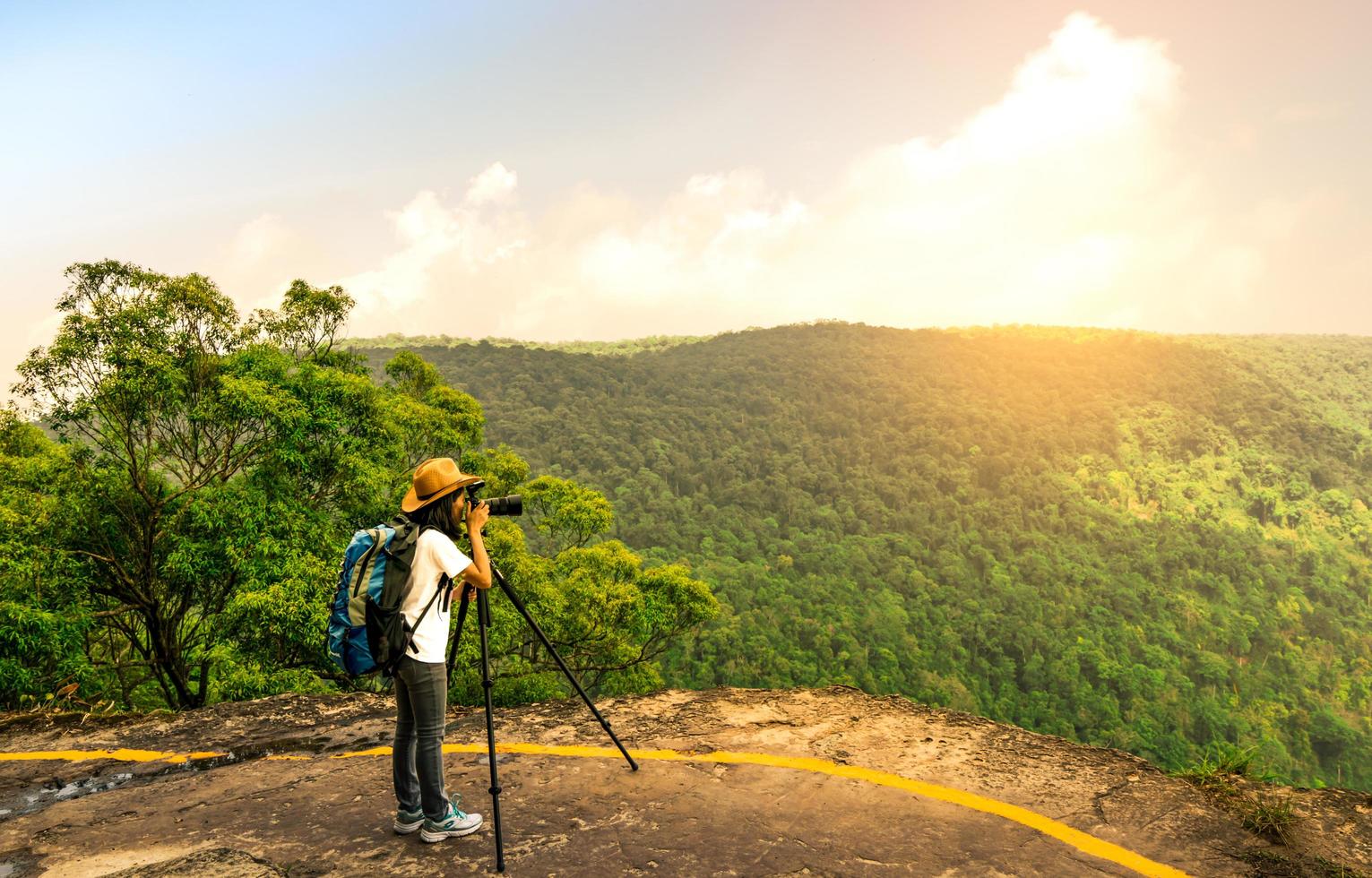 jonge reizende vrouw met rugzak hoed en camera op statief staan op de top van de berg klif kijken prachtig uitzicht op bossen en lucht op haar vakantie. Aziatische vrouw reist alleen. foto