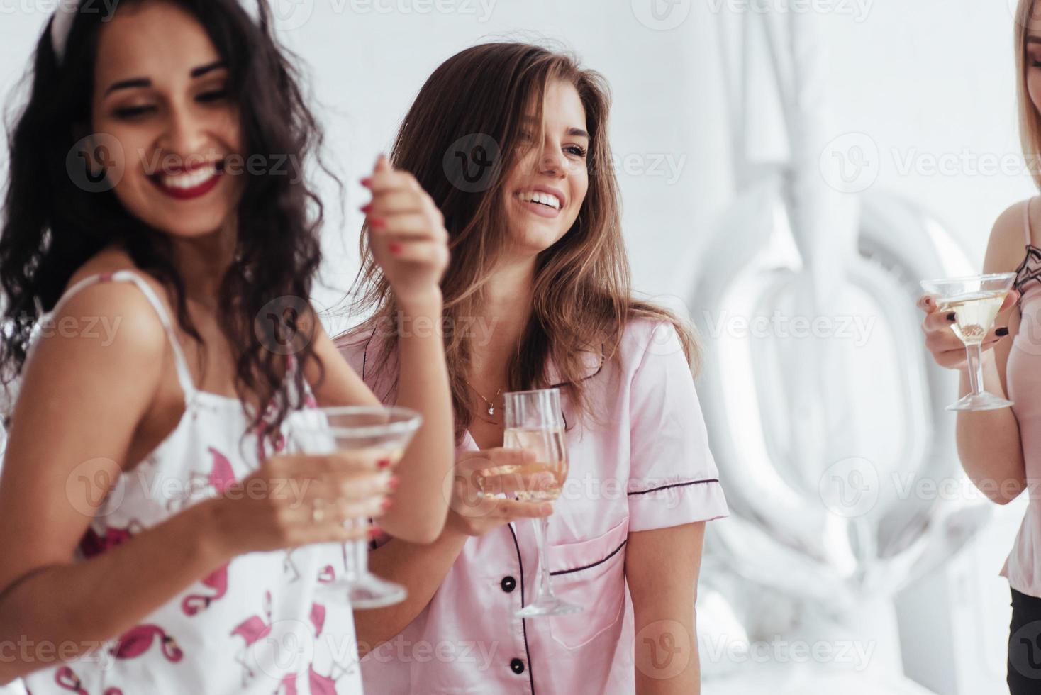 vrolijke dag. vrijgezellenfeest vieren in witte slaapkamer. meisjes glimlachen en drinken champagne foto