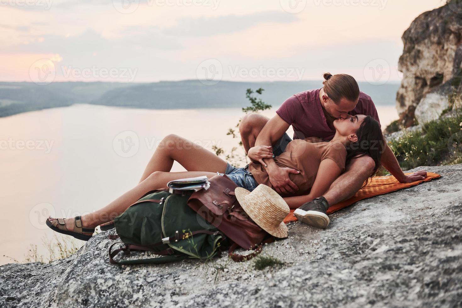 knuffelend paar met rugzak zittend op de top van rock mountain genieten van uitzicht kust een rivier of meer. reizen langs bergen en kust, vrijheid en actief levensstijlconcept foto