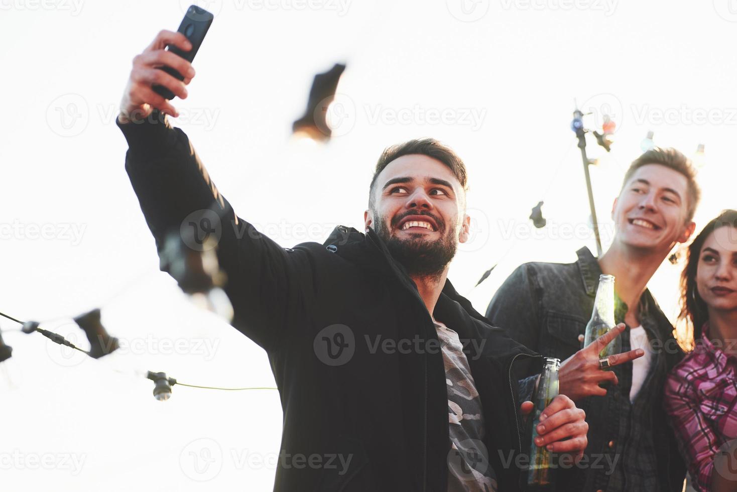 aantrekkelijke bebaarde man houdt de telefoon vast. groep jonge vrolijke vrienden die plezier hebben terwijl ze selfie op het dak nemen met versierde gloeilampen foto