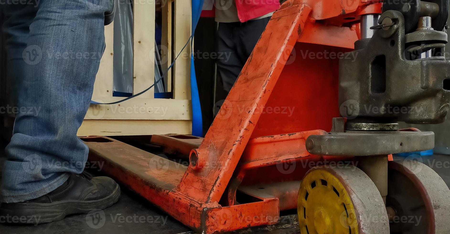 vorkheftruckstapelaar die houten kist van fabrieksmachine in magazijn behandelt. arbeider met veiligheidsschoenen die met handpalletstapelaar werken. goederen vervoer. magazijn en logistiek bedrijf. oranje palletwagen. foto