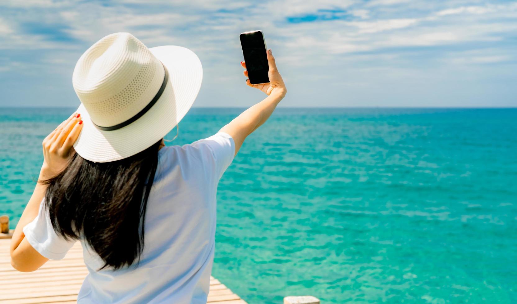 jonge aziatische vrouw draagt een strohoed in een casual stijl, gebruikt een smartphone die selfie neemt op een houten pier. zomervakantie op tropisch paradijsstrand. gelukkig meisje reizen op vakantie. vrouw genieten en ontspannen leven. foto