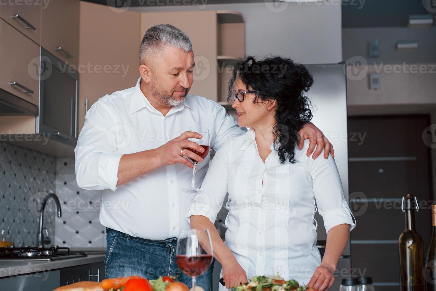 laten we wat wijn drinken. senior man en zijn vrouw in wit overhemd hebben een romantisch diner in de keuken foto