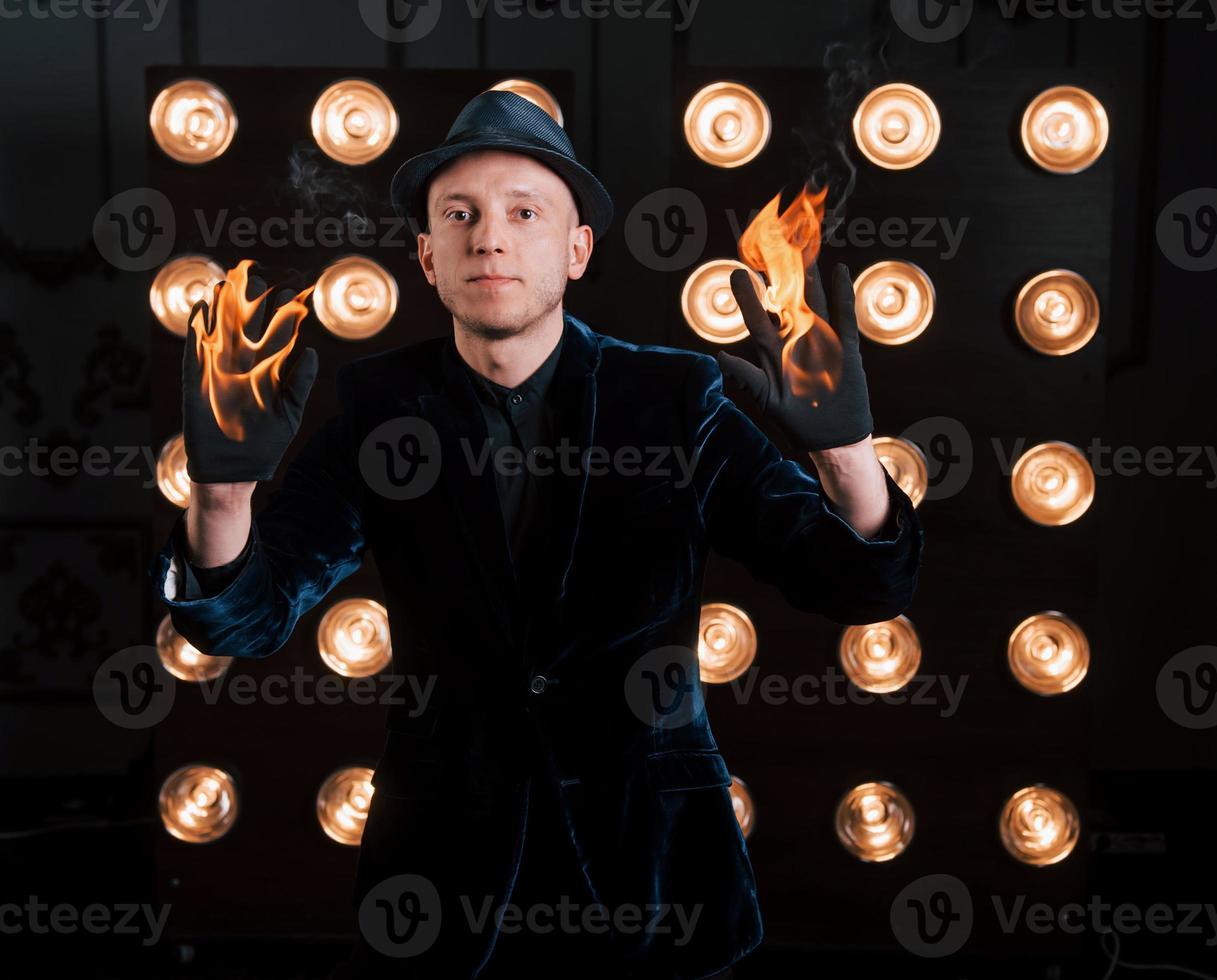 handen branden, maar dit is niet erg voor deze goochelaar. professionele illusionist in zwarte hoed, shirt en handschoenen spelen met vuur foto