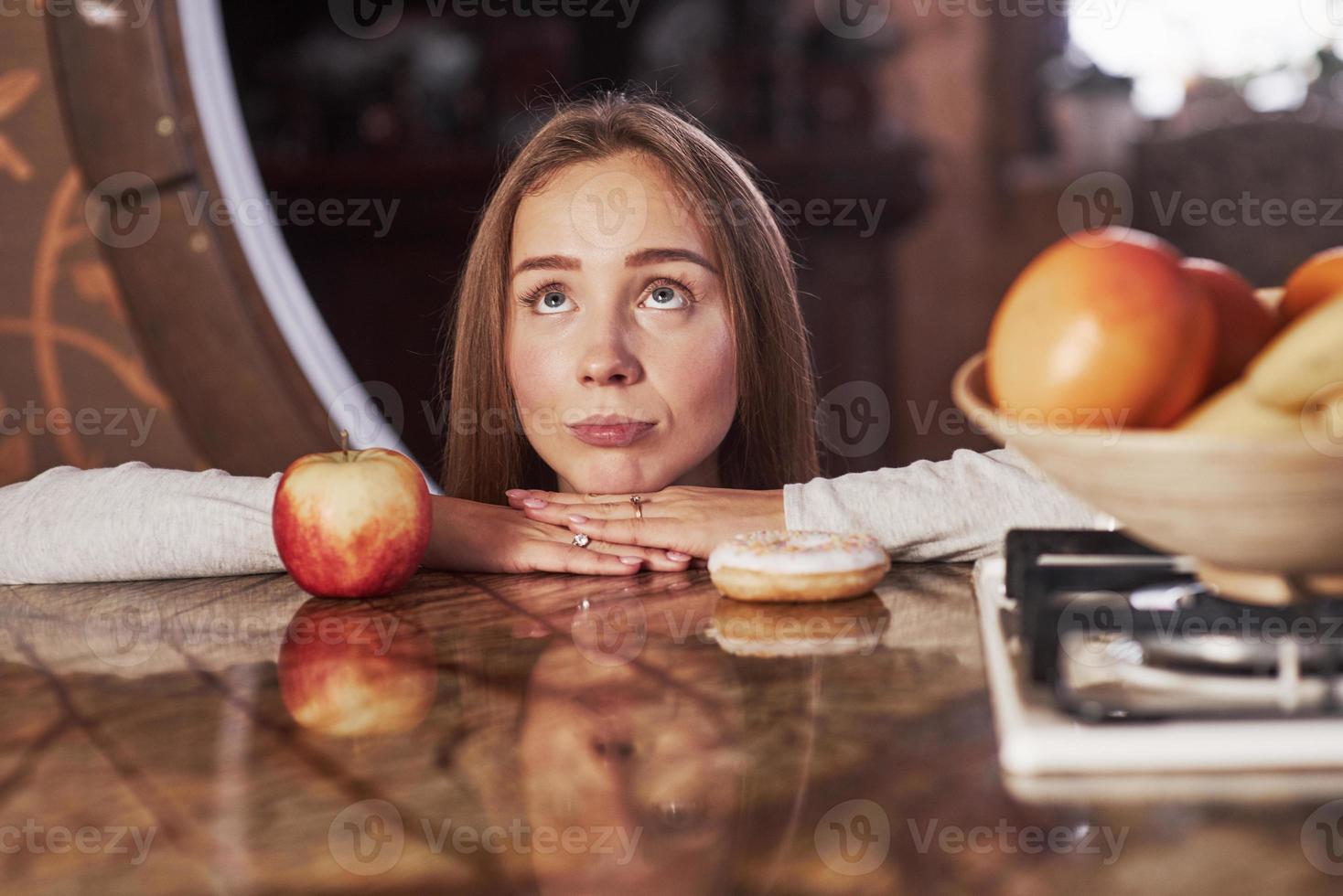 grappig gezicht maken. schattige jonge huisvrouw leunend op de tafel met appel en koekje en kijkt op foto