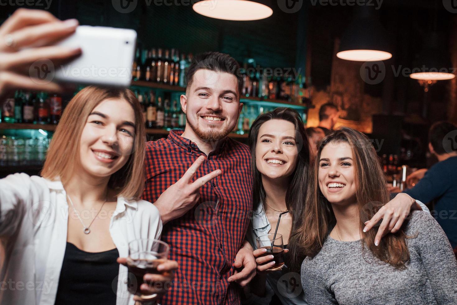 man toont gebaar van twee vingers. vrienden nemen selfie in prachtige nachtclub. met drankjes in de handen foto