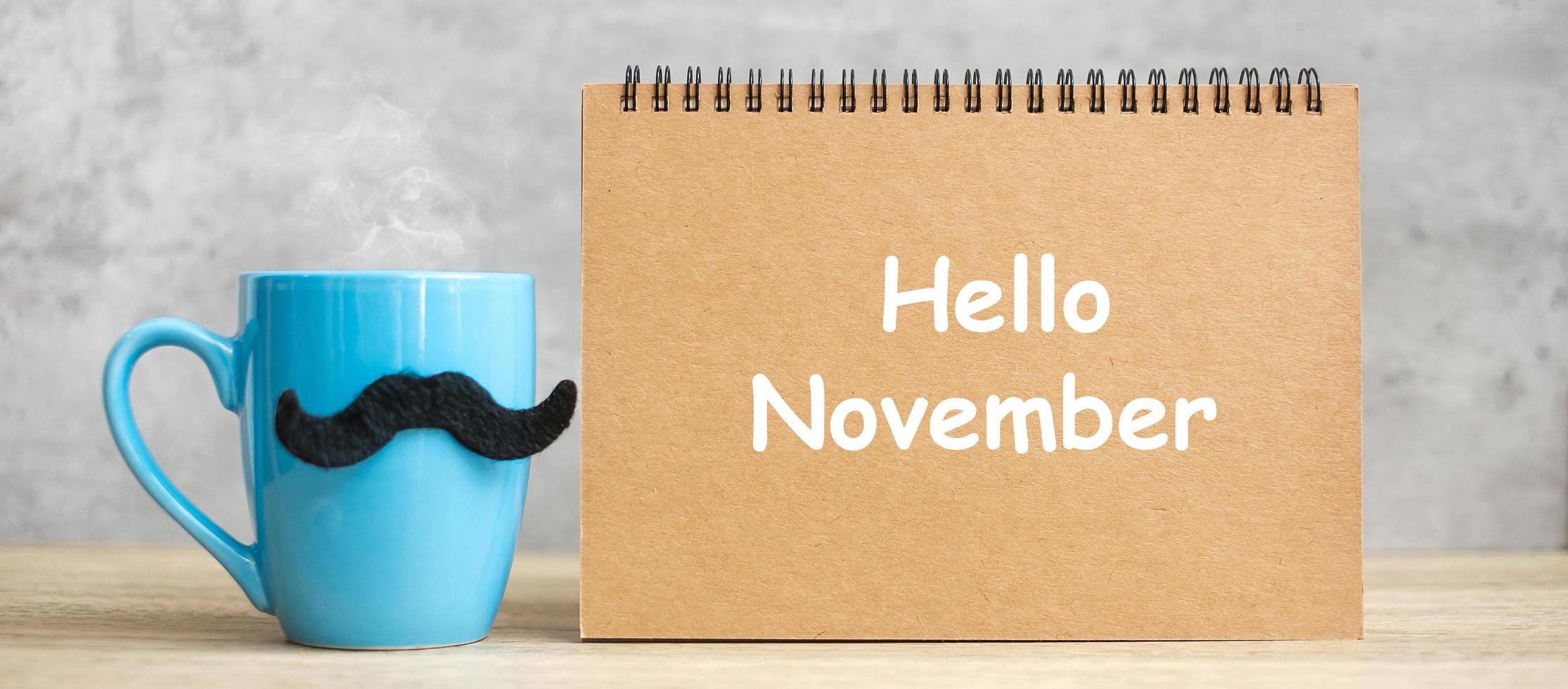 hallo november met papieren notitieblok, blauwe koffiekop of theemok en zwart snordecor op tafel. internationale mannendag, gelukkige vaderdag en feestconcept foto