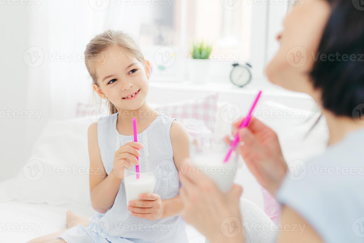 bijgesneden opname van mooi klein vrouwelijk kind drinkt smakelijke milkshake samen met haar moeder, geniet van goedemorgen en rustige huiselijke sfeer, heeft een gelukkige blik. mensen, ontbijt en beddengoed foto