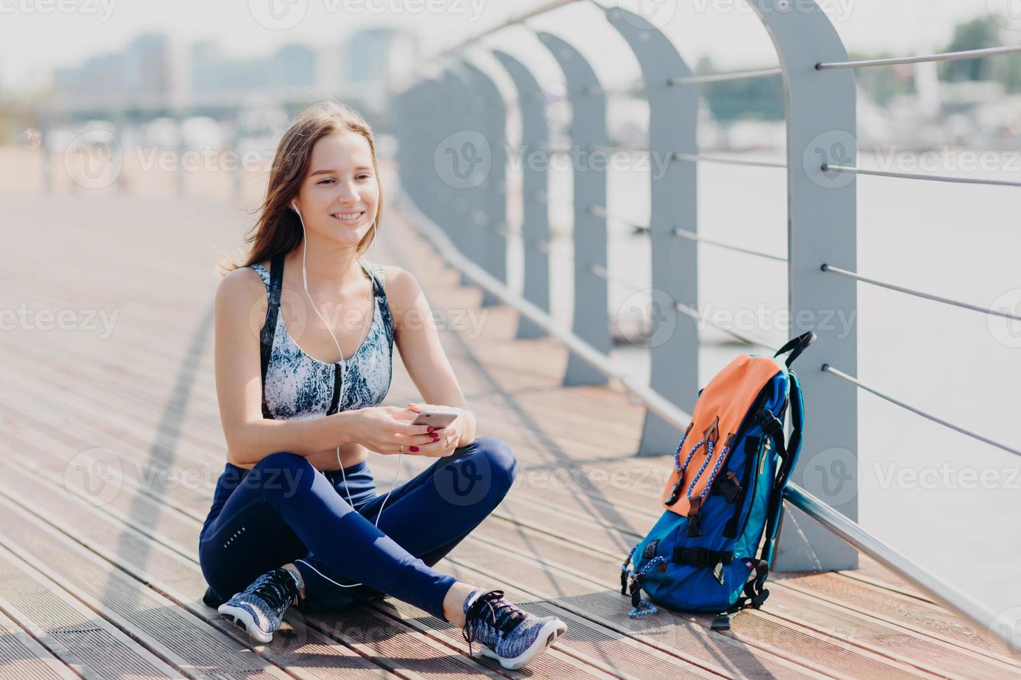 foto van een ontspannen vrolijk fitnessmeisje in sporswear, zit met gekruiste benen buiten, rust na een lange wandeling, luistert naar muziek met mobiel en koptelefoon, rugzak in de buurt. mensen, technologie en recreatie