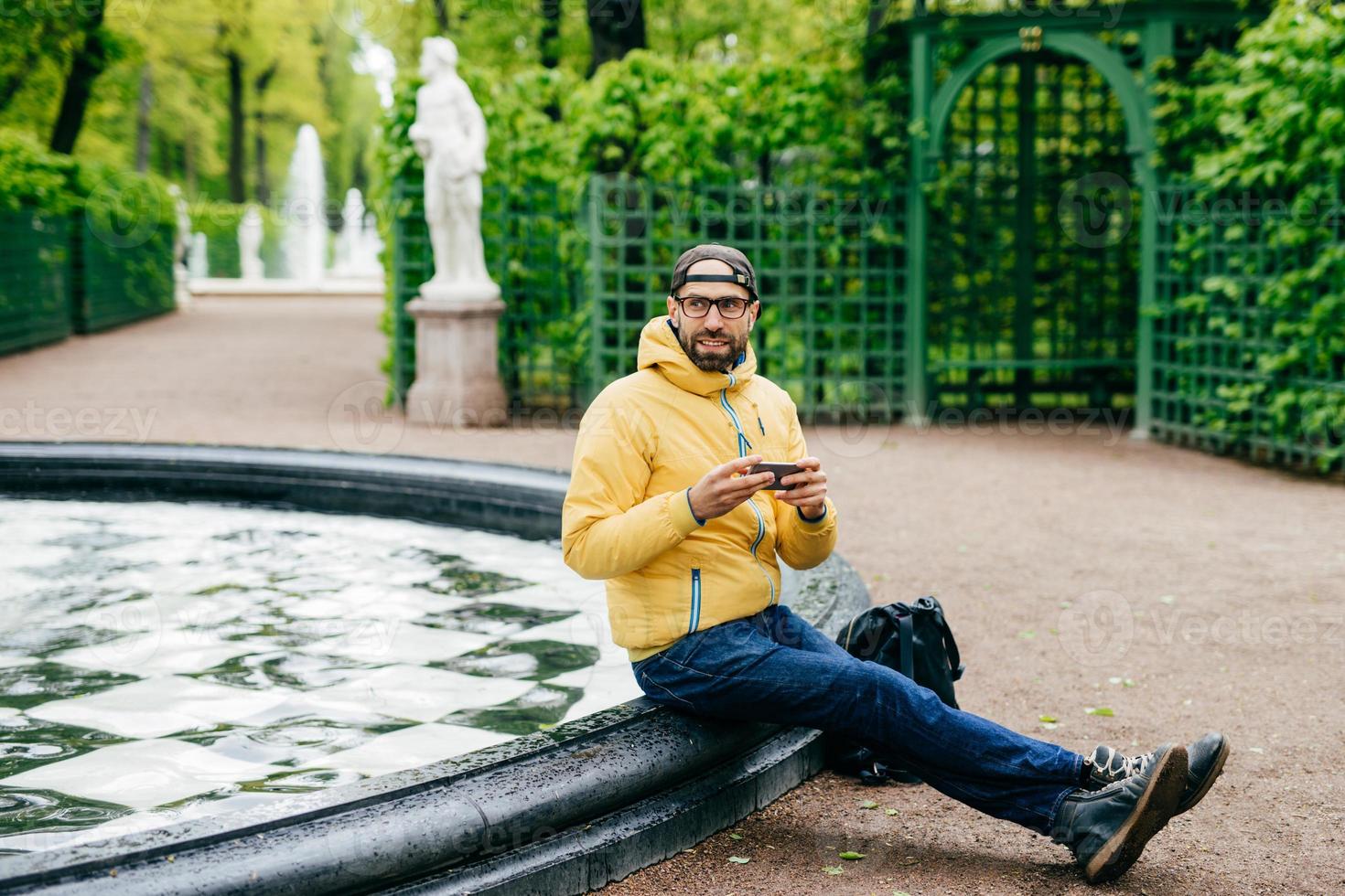 buitenportret van een bossige man met een pet, jas, spijkerbroek en bril met rust in de buurt van een fontein die zijn moderne apparaat in handen houdt en blij is zijn vriend op te merken. ontspannen hipster man in de open lucht foto