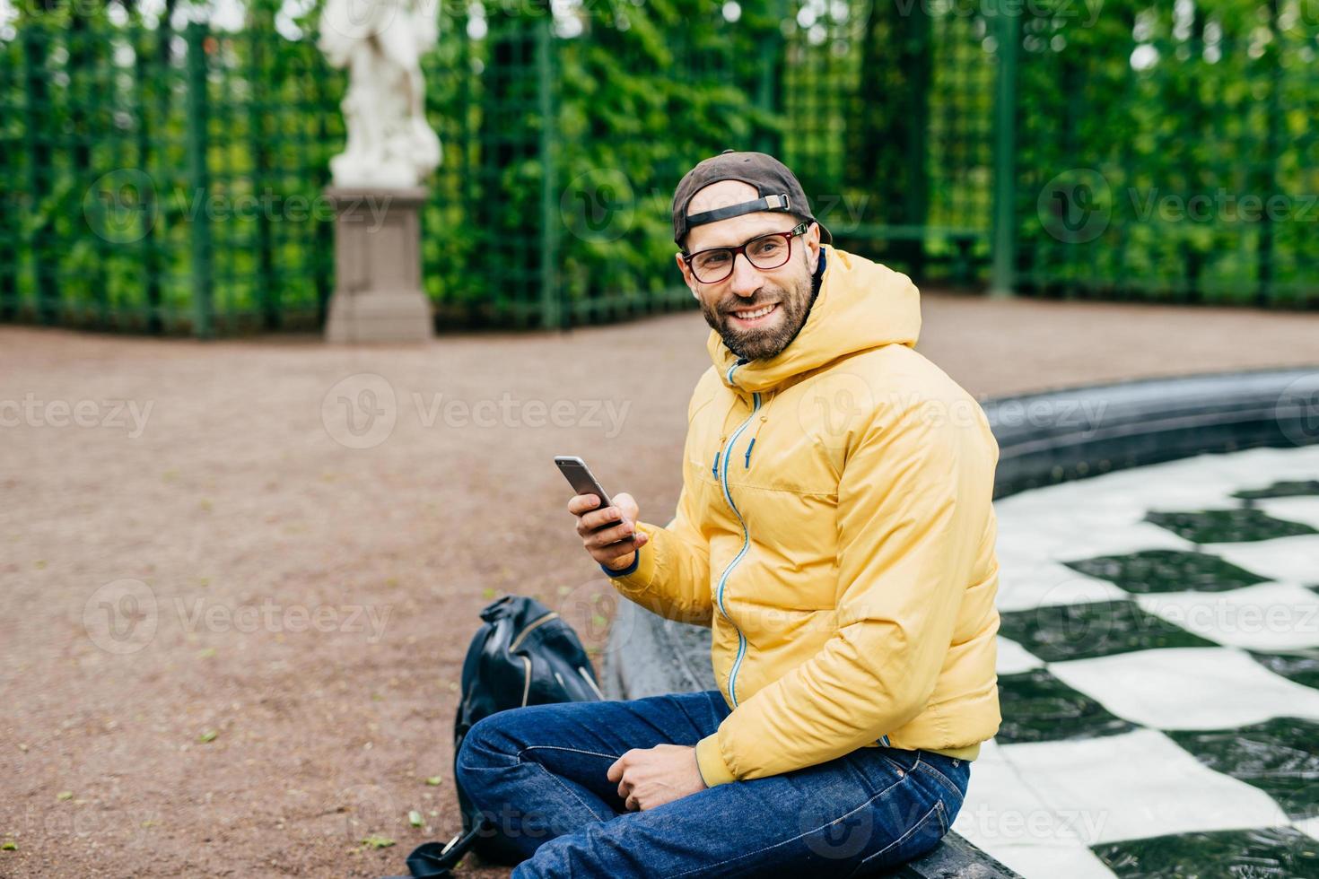 horizontaal portret van een bebaarde hipster-man die opzij kijkt met een heerlijke uitdrukking terwijl hij in de buurt van de fontein zit en foto's maakt van de prachtige natuur. stijlvolle man die buiten rust met mobiele telefoon foto