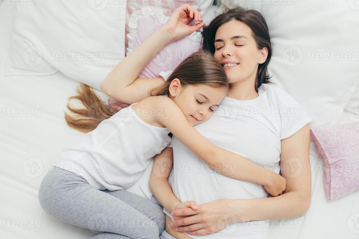 bovenaanzicht van vrolijke moeder slaapt op wit bed in de buurt van haar dochter die moeder met veel liefde omhelst, een goede houding toont, prettige dromen heeft en lekker rust, een pyjama draagt. mensen, slaapconcept foto