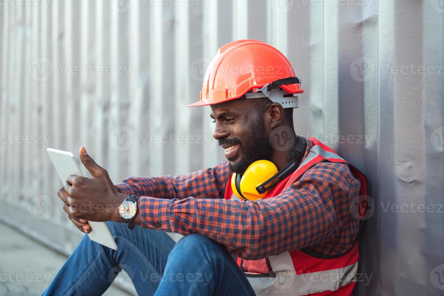 close-up gelukkig lachende mannelijke Afro-Amerikaanse ingenieur met veiligheidsvest en helm, zittend en met behulp van mobiele telefoon op logistieke vrachtcontainers werf. foto