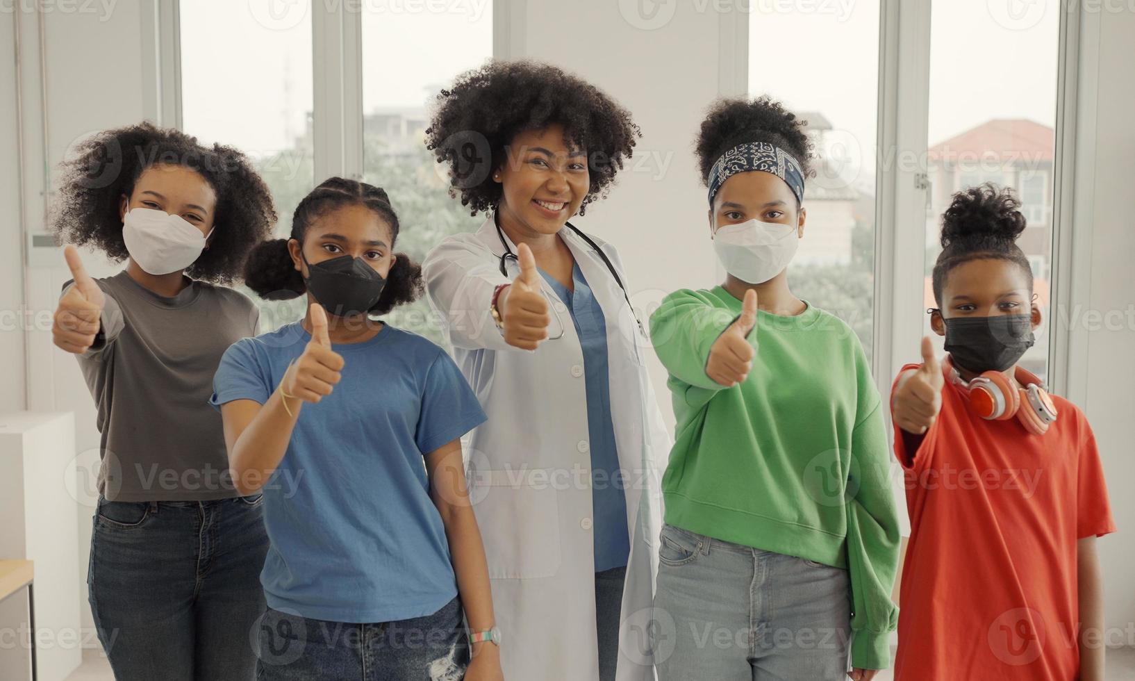 Afro-Amerikaanse arts en kinderen dragen een masker, gelukkig duimen omhoog vinger in het ziekenhuis of de kliniek. foto