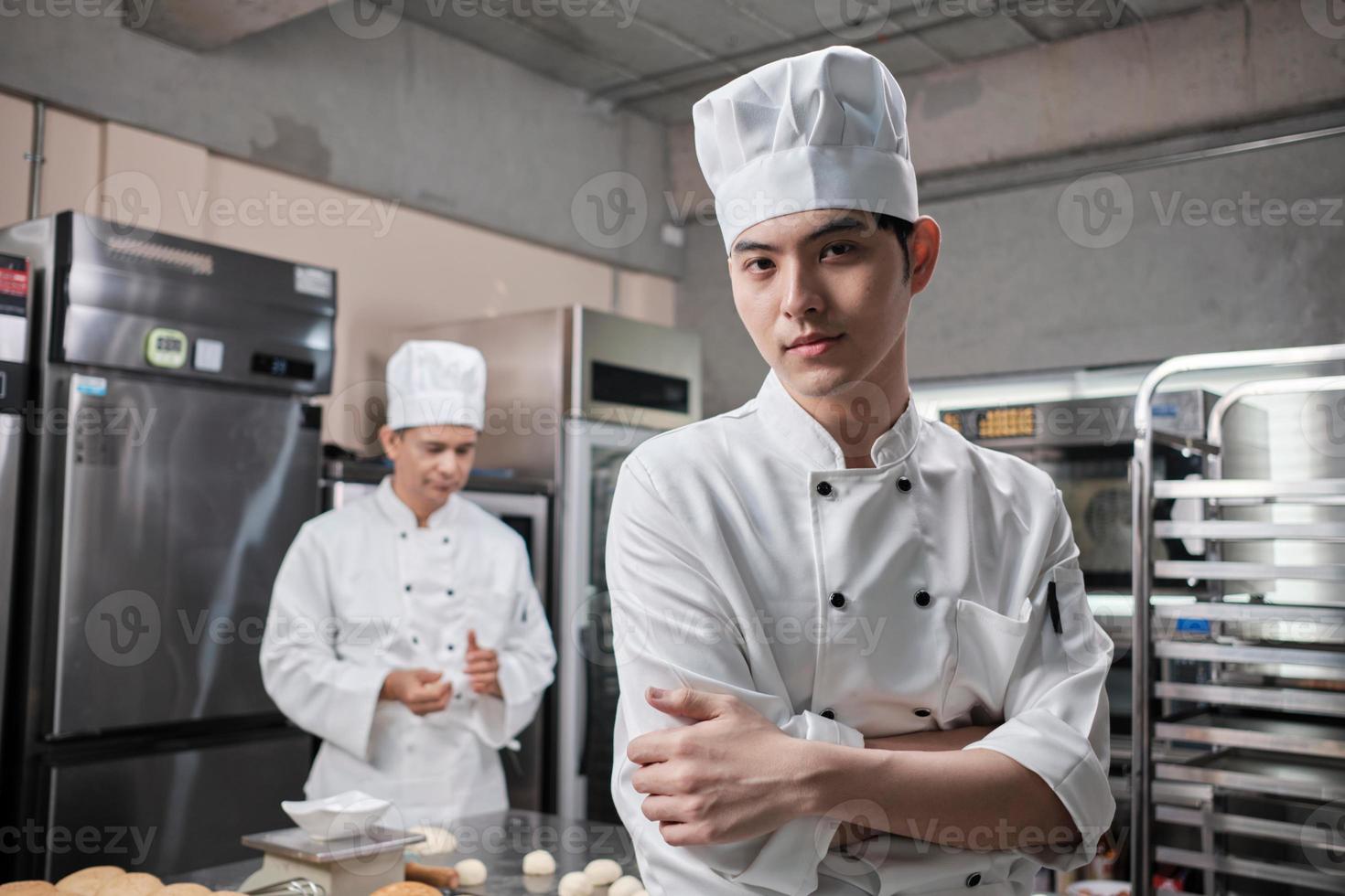 portret van een jonge Aziatische mannelijke chef-kok in wit kookuniform kijkend naar de camera, armen gekruist met vertrouwen, voedingsmiddelen professionele bezetting, commerciële gebak culinaire banen in de keuken van een restaurant. foto