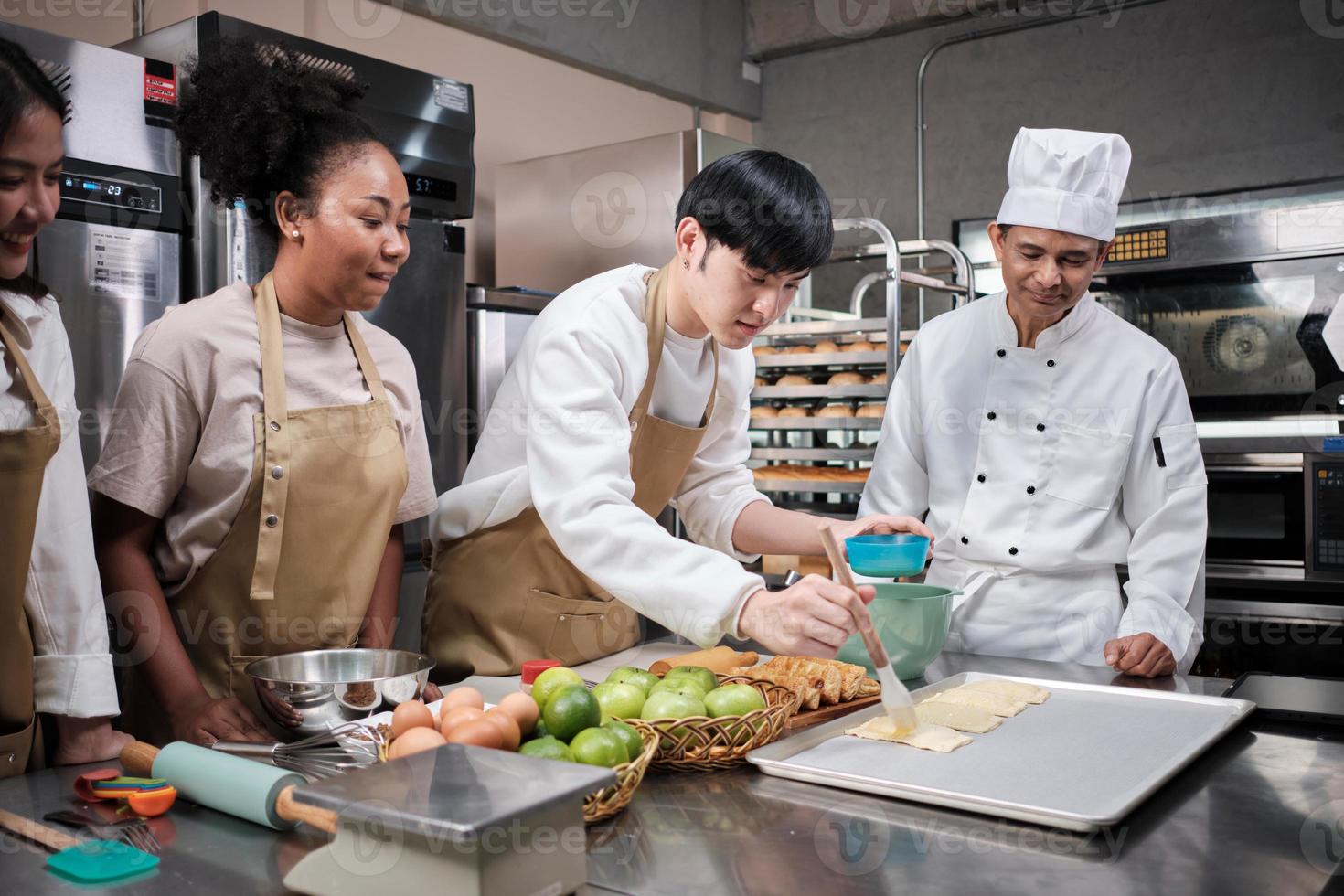 keukencursus, senior mannelijke chef-kok in uniform leert jonge kooklesstudenten, borstelt gebakdeeg met eierroom, bereidt ingrediënten voor bakkerijproducten, fruittaarten in roestvrijstalen keuken. foto