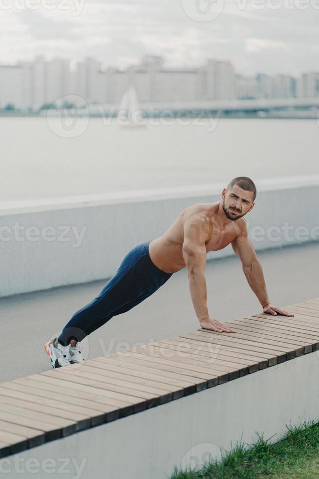 buitenopname van vastberaden gespierde man warmt zich op voor de training staat in plankhouding heeft naakte torso gespierd lichaam geniet van trainen in de open lucht draagt actieve kleding heeft training om vitaliteit te behouden foto