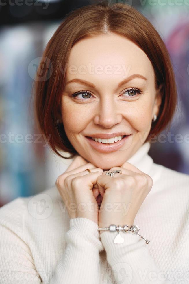 foto van aantrekkelijke jonge vrouw met bruin steil haar, kijkt vrolijk weg, gekleed in witte kleding, draagt witte coltrui, houdt handen onder de kin, drukt vreugde uit, heeft vriendelijke uitstraling