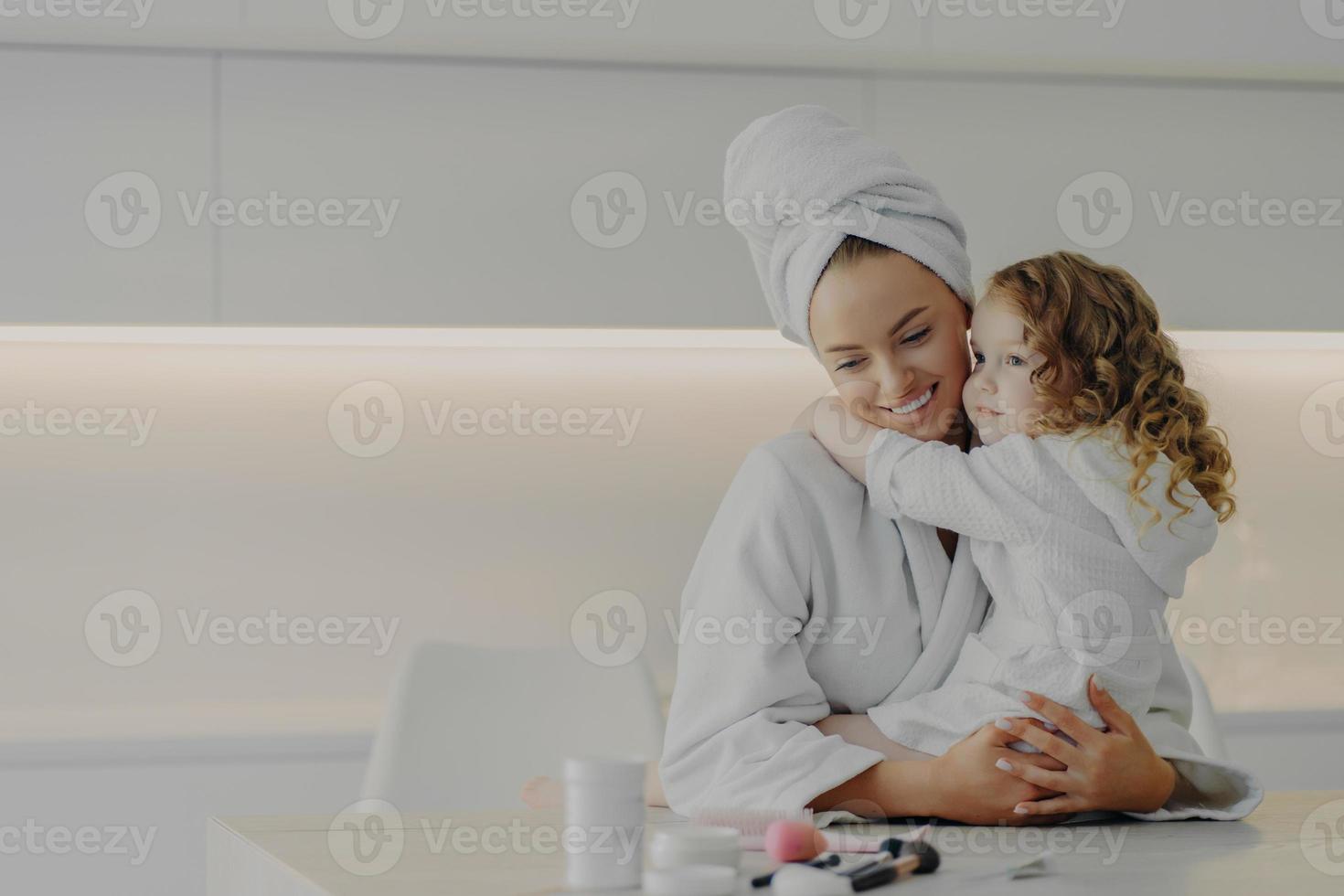 moeder en schattig dochtertje in witte badjassen knuffelen en genieten van het weekend samen thuis foto