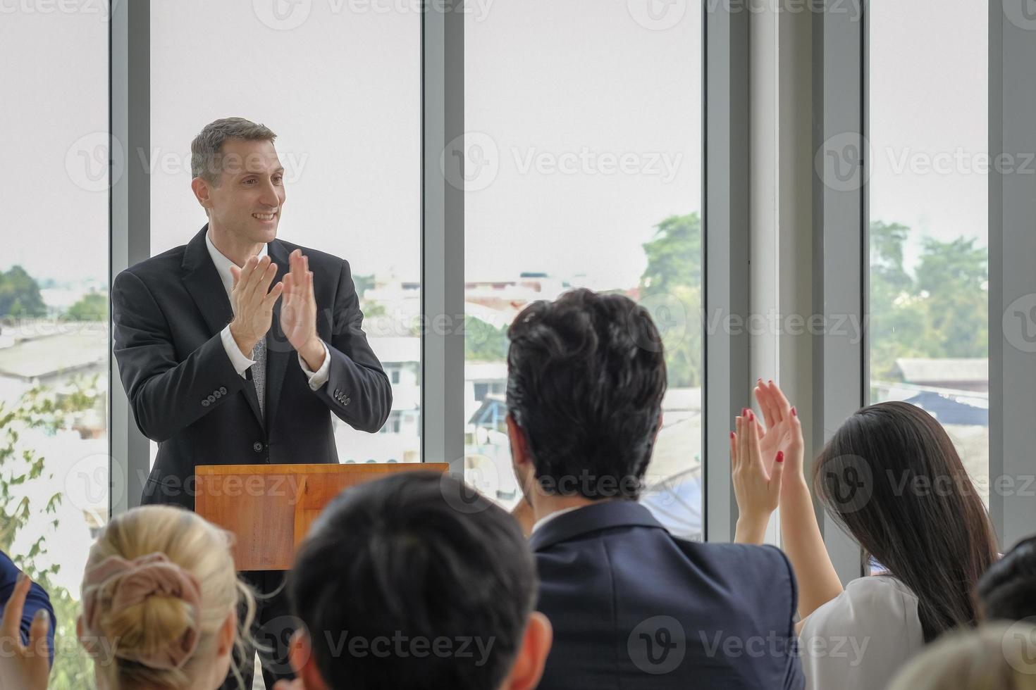 gelukkige kaukasische zakenman staat in de handen te klappen op het podium om te vieren met andere deelnemers aan het seminar-evenement. selectieve aandacht. foto