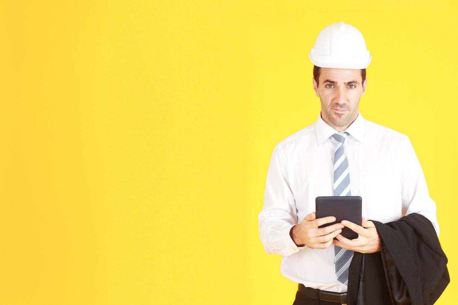 knappe en slimme ingenieur in pak en wit overhemd en het dragen van een witte veiligheidsingenieur hoed met hand met smartphone geïsoleerd op gele achtergrond. kopieer ruimte foto