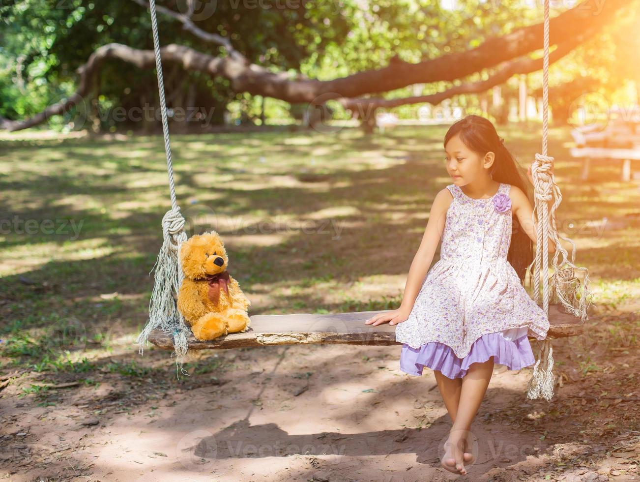 schattig klein meisje zit schommels, teddybeer zit met haar, klein meisje wees naar de boom. foto