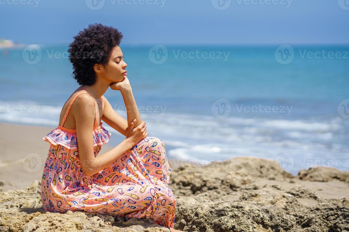 jonge zwarte vrouw zittend op rotsen op het strand, ogen gesloten, ontspannen met mindfulness-technieken. foto