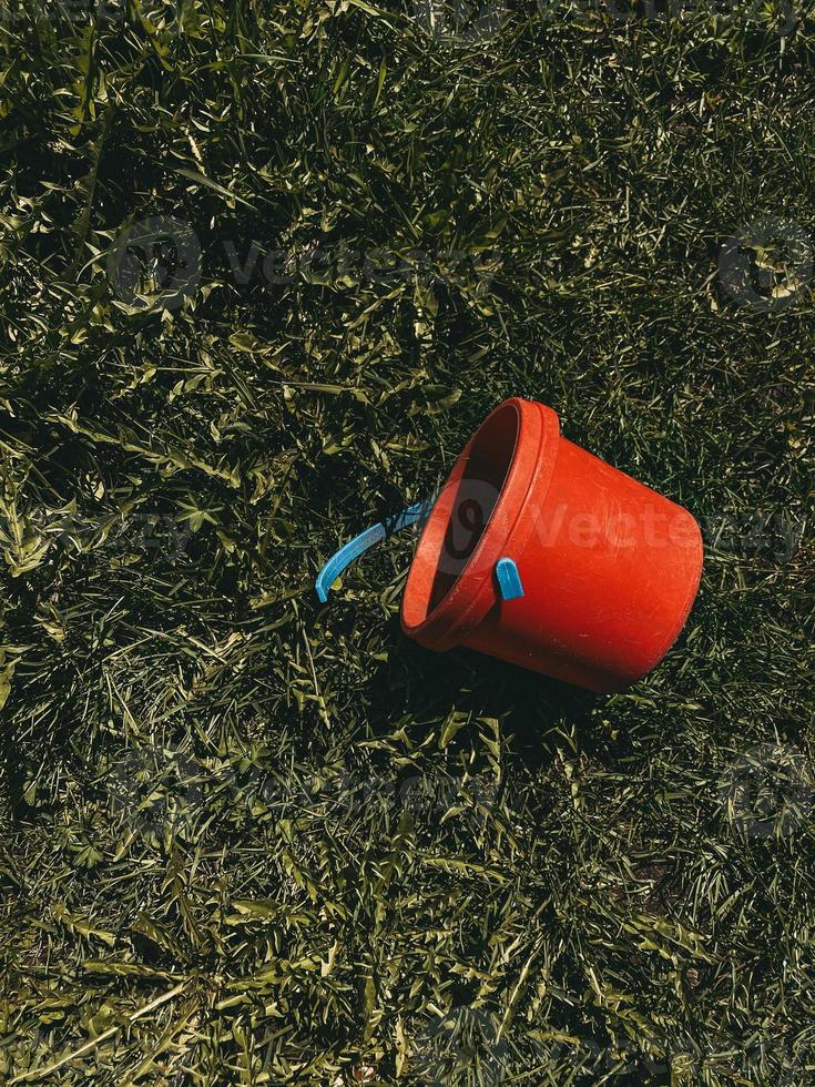 rode speelgoedemmer met gebroken blauw handvat liggend op gras, verticale foto, warme kleuren. plastic kinderspeelgoed om met zand of water te spelen, tuingereedschap; foto