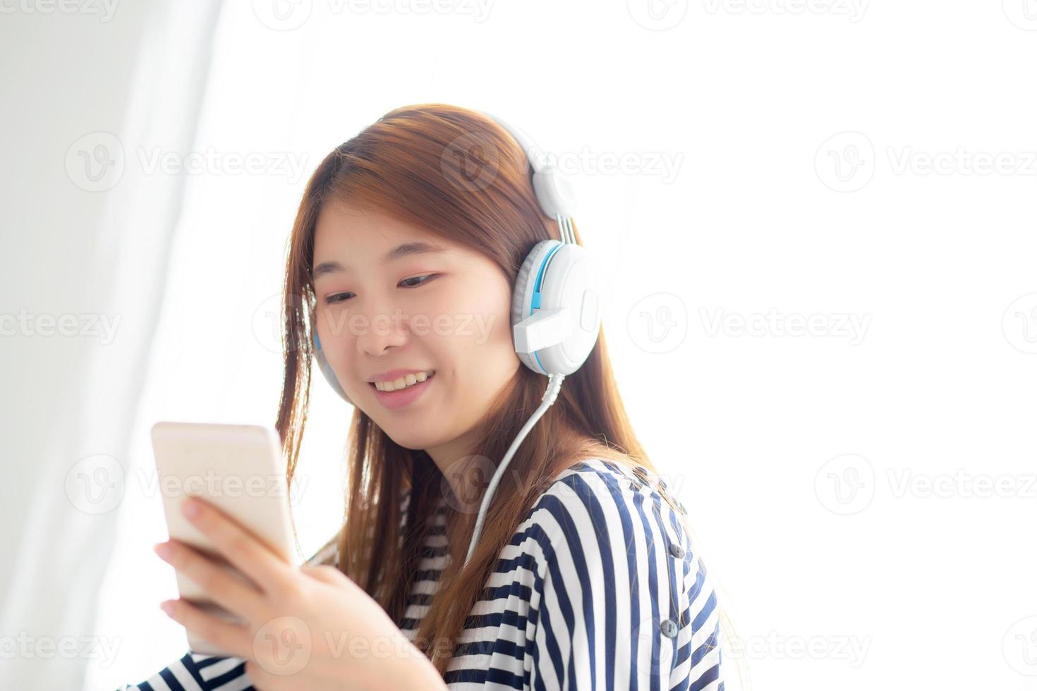 mooie aziatische jonge vrouw geniet van luister muziek met hoofdtelefoon en houdt slimme mobiele telefoon vast terwijl ze in de slaapkamer zit, ontspan meisje met oortelefoon, vrije tijd en technologie concept. foto