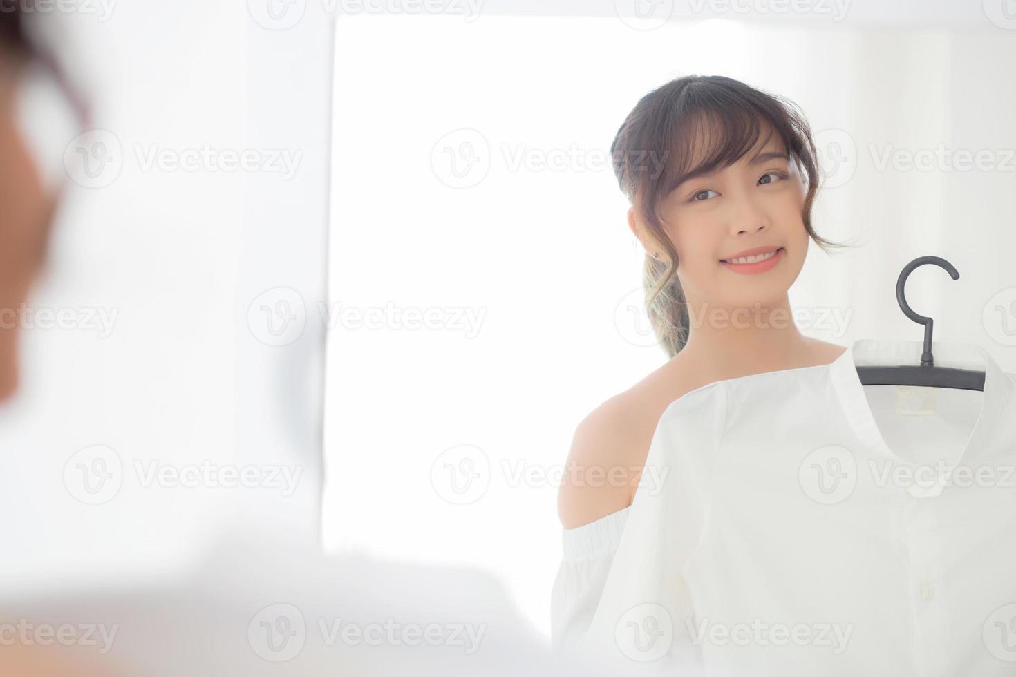 jonge aziatische vrouw die lacht met kleding die zich aankleedt met een moderne en uitziende reflectiespiegel in de kamer, meisje kiest kledingmode met een gelukkig en vrolijk, lifestyle-concept. foto