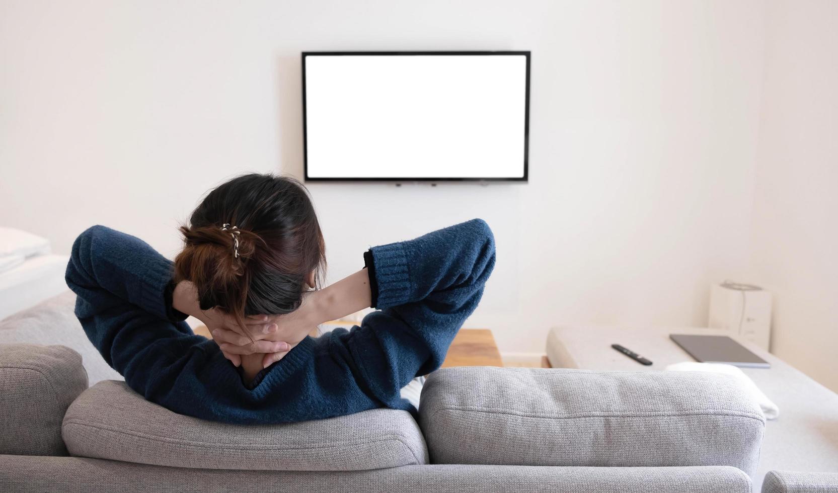 achter van een aziatische jonge vrouw die thuis op de bank op een wit scherm tv kijkt foto