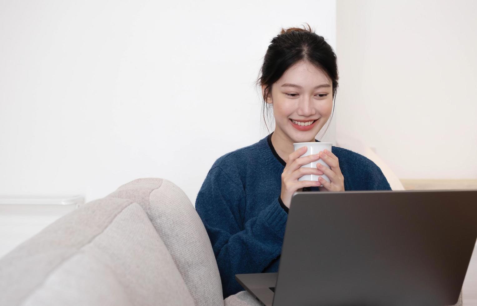 portret mooie jonge aziatische vrouw die op laptop online werkt, internet gebruikt en de beker vasthoudt. thuis op de bank zitten, vrije ruimte foto