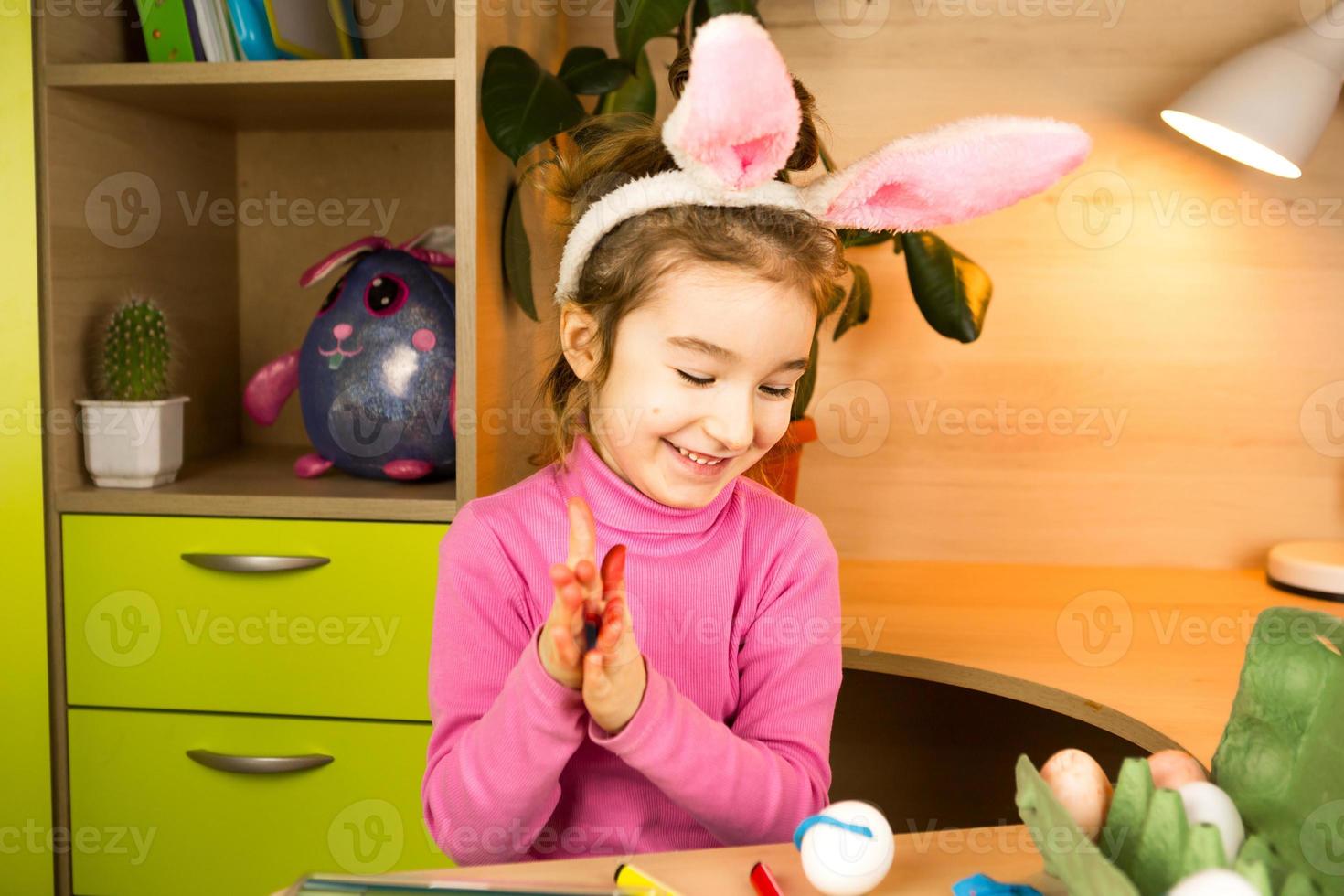 een meisje in de oren van een paashaas schildert eieren met een viltstift in het interieur. knutselen, voorbereiding voor een religieuze feestdag, een dienblad met eieren, hazenoren gemaakt van plasticine foto