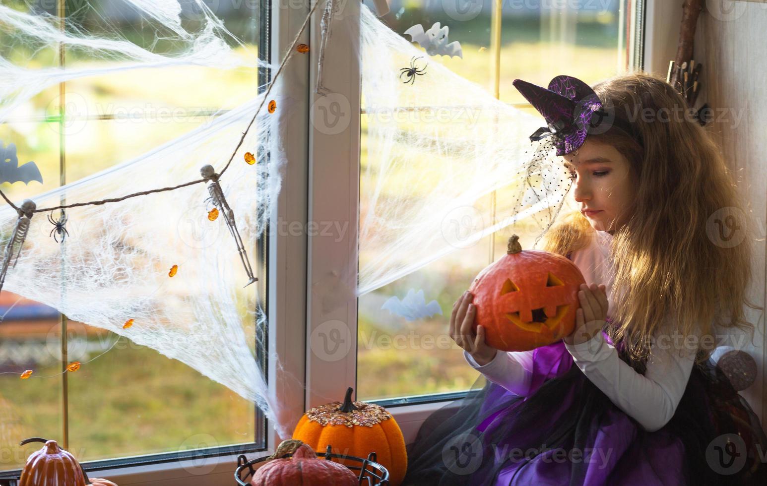 een meisje in een heksenkostuum op de vensterbank bij het raam op een halloween-feest, het huis spinnenwebben, vleermuizen, spinnen, pompoenen. verschrikkelijk landschap, angst en horror foto