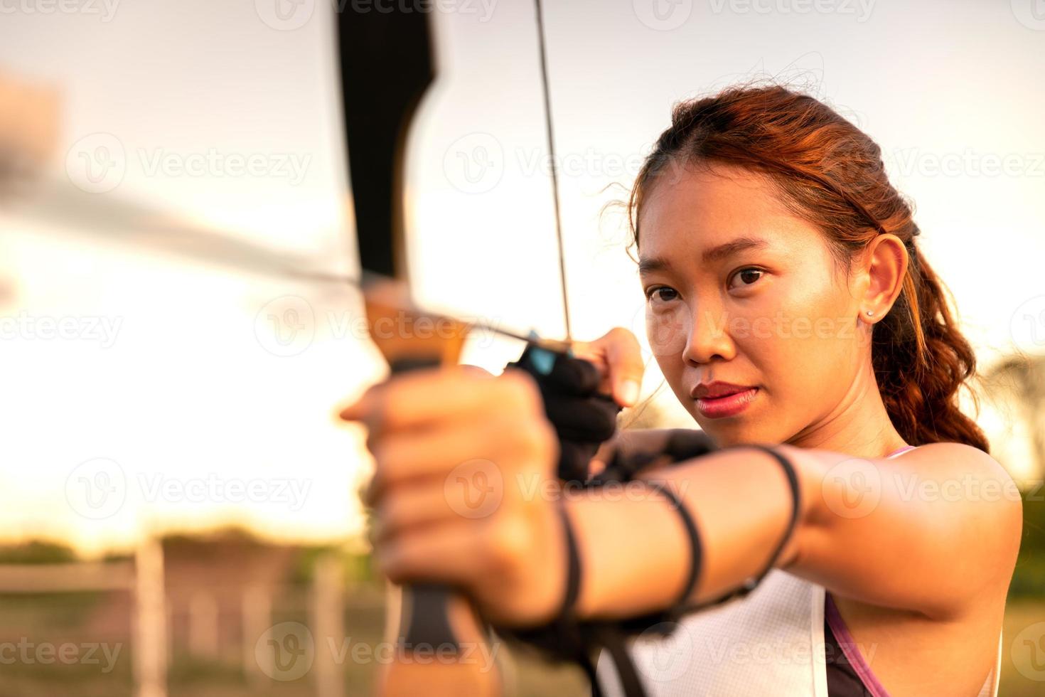 jonge vrouwelijke boogschutter, boogschieten, schiet met boog in het natuurveld om te richten, succesconcept, op het veld voor sportoefening bij zonsondergang foto